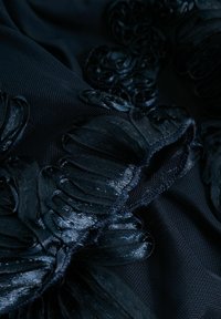 Gros plan sur un tissu noir avec une broderie florale texturée et des pétales de satin superposées créant un effet 3D sur un fond en maille.