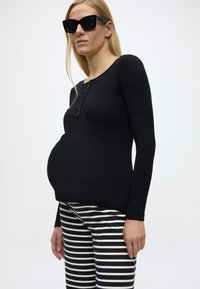 Έγκυος γυναίκα που φοράει μαύρο μακρυμάνικο τοπ, μαύρα γυαλιά ηλίου και μαύρο-άσπρο ριγέ παντελόνι, στέκεται μπροστά σε απλό φόντο.