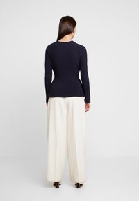 Ribbineulakattu merinonsininen pusero, jossa on istuva vyötärö ja liehuva helma, yhdistettynä kermaisiin leveälahkeisiin housuihin, jotka ovat yksiväriset ja minimalistisen malliset.