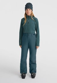 Tealfärgad fleece-tröja med halvdragkedja och logotyp, ihop med tealfärgade skidbyxor med hög midja och svarta detaljer. Svarta skidstövlar syns nedanför.