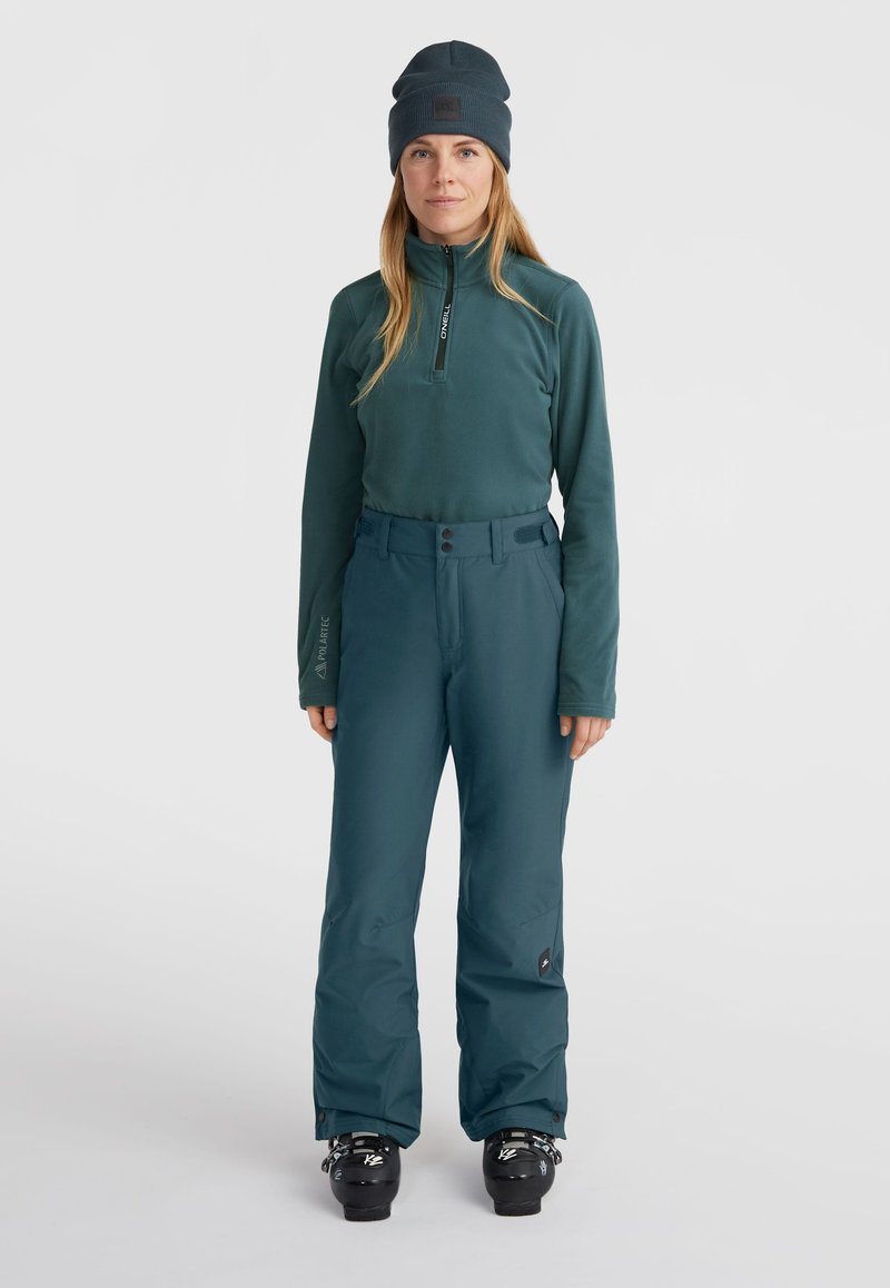 Teal fleece half-zip trui met logo, gecombineerd met teal skibroek met een hoge tailleband en zwarte hardware. Zwarte skischoenen zijn zichtbaar onderaan.