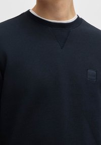 Námořnicky modrá mikina s kulatým výstřihem, texturovanou látkou a malou černou čtvercovou nášivkou s logem „BOSS“ na hrudi.