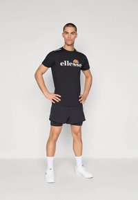 Must sportlik t-särk "ellesse" logoga, mustade pükstega. Modell kannab valgeid tossusid ja pahkluu pikkusega sokke. Üldmulje on minimalistlik.