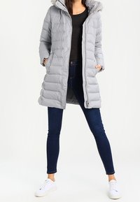 Vaaleanharmaa untuvatakki, jossa on huppu, tikattu muotoilu ja sivutaskut, päällä musta paita ja tummat farkut, yhdistettynä valkoisiin tennareihin.