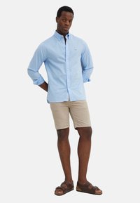 Camisa de boton azul claro, pantalones cortos beige y sandalias marrones. La camisa tiene un cuello clásico y las mangas arremangadas.