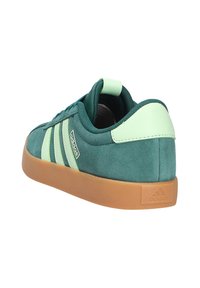 Zapatillas de gamuza verde con acentos en verde claro y logo de tres rayas. Suela de goma marrón con una superficie texturizada para mayor agarre.