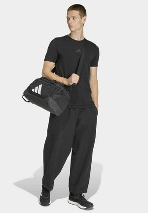 Borsa da palestra atletica nera con strisce diagonali bianche, indossata con una maglietta nera a maniche corte e pantaloni neri larghi, su sfondo bianco.