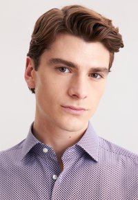 Camicia bianca con un piccolo motivo geometrico bordeaux, colletto con bottoni e tessuto morbido. Modello mostrato dalla spalla in su.