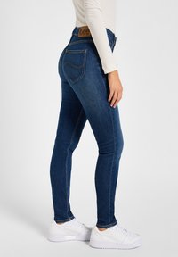 Jeans skinny en denim bleu avec une taille mi-haute, featuring une étiquette en cuir marron, des poches arrière et une coupe slim jusqu'à la cheville.