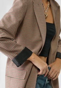 Blazer a cuadros marrones y rojos con un diseño estructurado, que cuenta con puños de satén negro y un único bolsillo frontal, combinado con un top corto negro.