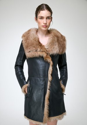 Femme portant un manteau en cuir noir avec un grand col et bordure en fourrure beige, mains dans les poches, debout devant un fond clair uni.