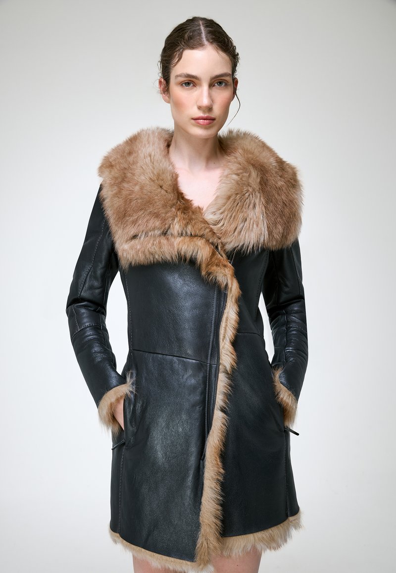 Femme portant un manteau en cuir noir avec un grand col et bordure en fourrure beige, mains dans les poches, debout devant un fond clair uni.