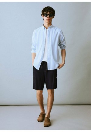 Ljusblå randig skjorta med knappar, vit t-shirt, svarta cargoshorts, bruna mockaskor och solglasögon i mörkt på en modell mot en grå bakgrund.