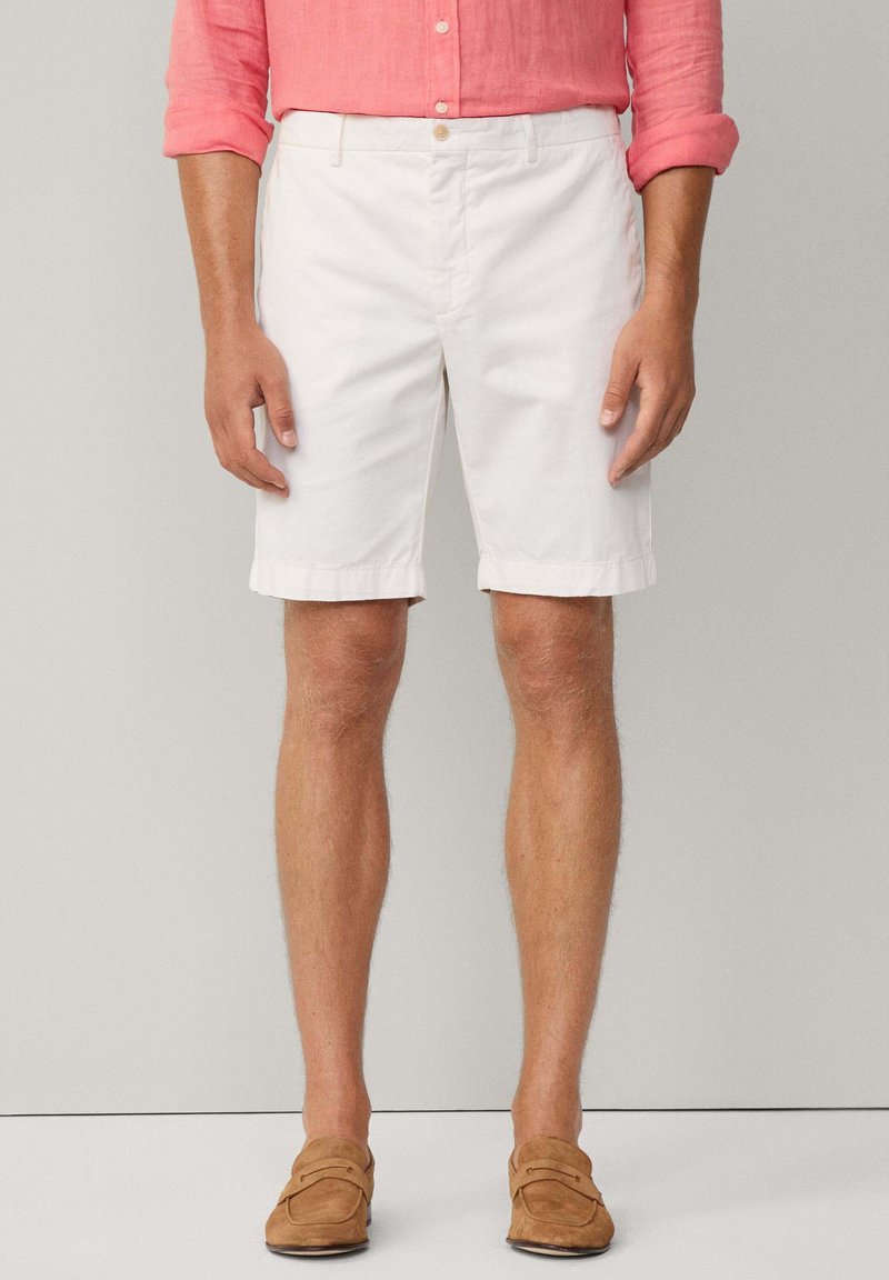 Shorts bianchi in cotone con fronte piatto, orlo piegato e tasche laterali, abbinati a una camicia button-up rosa chiaro e scarpe marroni slip-on.