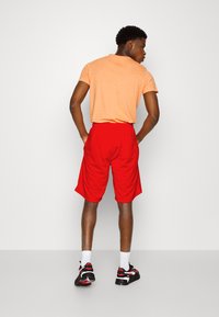 T-shirt arancione a maniche corte e pantaloni corti rossi. I pantaloni corti hanno tasche laterali, mentre il t-shirt presenta un colletto a girocollo. Scarpe da ginnastica nere e rosse indossate.