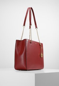 Punainen synteettinen kangaskassi, jossa on teksturoitu pinta, kultaiset ketjusomisteet ja koristeellinen riipus. Ominaisuuksiin kuuluu kaksi olkahihnaa ja tasainen pohja.