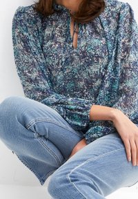 Blouse fluide à fleurs bleues avec un col haut et un détail noué, manches longues avec poignets froncés, associée à un jean bleu clair.