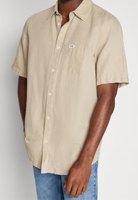 Uomo che indossa una camicia beige a maniche corte con taschino e jeans blu, in piedi con una mano rilassata al suo fianco.