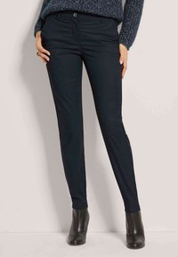 Marineblaue Slim-Fit-Hosen aus glattem Stoff, mit Knopfverschluss, Taschen und einem sauberen Saum. Kombiniert mit schwarzen, hochhackigen Stiefeletten.