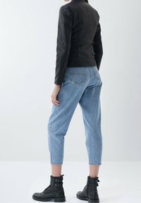 Persona che indossa una giacca di pelle nera, jeans a vita alta di colore blu chiaro e stivaletti neri, di spalle su uno sfondo neutro.