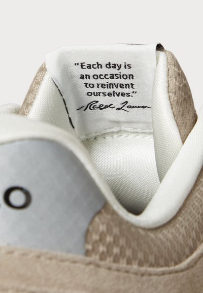 Bež tenisice s teksturiranom površinom na jeziku, srebrnim detaljima i bijelim postavom. Ima ispisanu poruku unutar cipela.