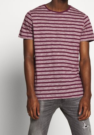 Mand iført en kortærmet T-shirt med bordeaux og grå striber og grå slidte jeans, stående mod en ensfarvet baggrund.