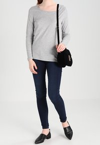 Graues Langarmoberteil, enganliegende dunkle Jeans und schwarze Slipper. Schwarze Schultertasche mit Klappe und kleiner dekorativer Hardware.