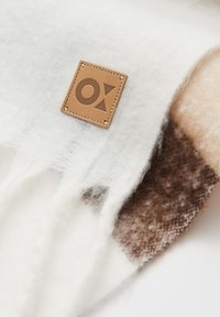 Weißer und brauner Wollmischschal mit einem Lederetikett mit eingeprägtem Logo, strukturierten Fransenrändern und einem weichen, warmen Aussehen.