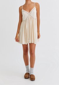 Cremfarbendes Kleid mit Spitzenakzenten, V-Ausschnitt und dünnen Trägern; verfügt über ein fließendes Design und gerafften Stoff an der Taille.