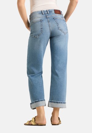 Vrouw met lichtblauwe opgerolde jeans, witte top en platte gouden sandalen, gezien van achteren tegen een effen achtergrond.