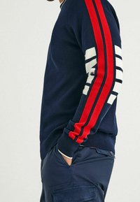 Suéter azul marino con acentos a rayas blancas y rojas en las mangas. Puños y cuello de canalé, textura suave, diseño ajustado.