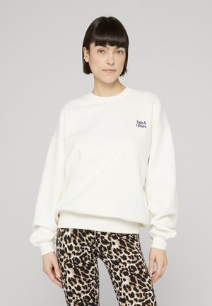 Kvinne med kort, mørkt hår iført en hvit genser med teksten "Salt & Olives" og leggings med leopardmønster, stående mot en ensfarget bakgrunn.