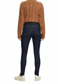 Suéter marrón de punto con diseño de trenzas, combinado con jeans skinny azul oscuro, que tienen bolsillos traseros y zapatillas blancas.