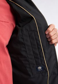 Gilet nero realizzato in tessuto liscio, con bordo piping, chiusura con zip e tasca interna. Accompagnato da una camicia rossa a maniche lunghe.