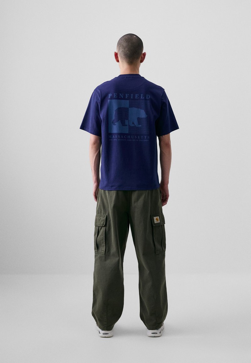 T-shirt marine avec un graphique d'ours et du texte au dos, associé à un pantalon cargo vert olive avec des poches latérales et un patch logo.