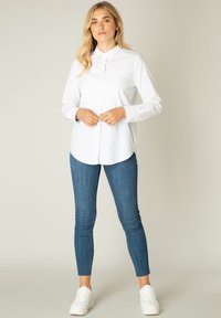 Witte overhemd met puntkraag, lange mouwen en een relaxed fit, gecombineerd met blauwe skinny jeans en witte sneakers.