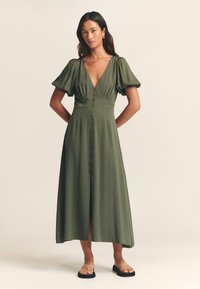 Robe midi vert olive avec des manches bouffantes, décolleté en V profond, boutonnière à l'avant et fente haute, associée à des sandales noires plates.