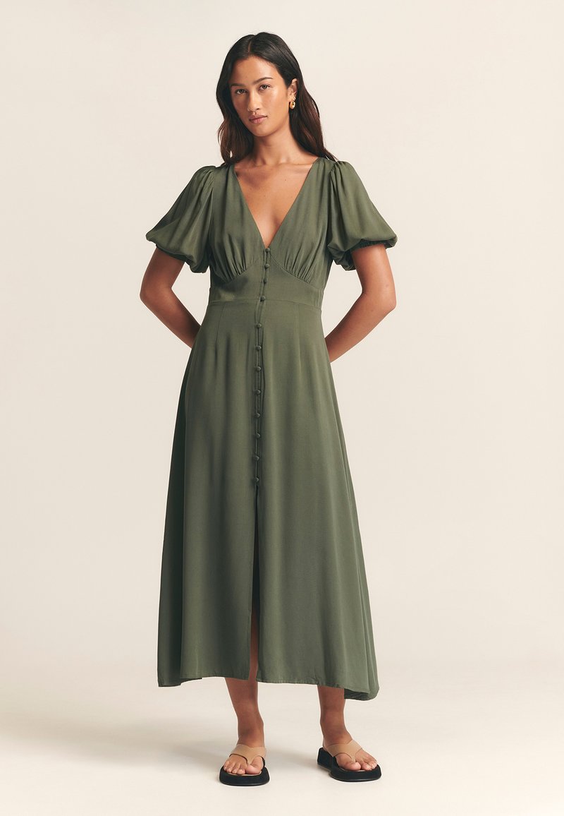 Robe midi vert olive avec des manches bouffantes, décolleté en V profond, boutonnière à l'avant et fente haute, associée à des sandales noires plates.