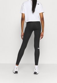 Leggings noirs avec une texture lisse, un design ajusté et un logo blanc près de la cheville. Associés à un T-shirt blanc court et des chaussures de sport.