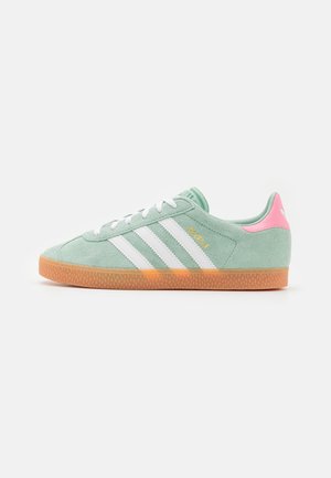 Zapatilla Adidas de ante verde claro con rayas blancas, suela de goma, lengüeta trasera rosa y texto dorado "Gazelle" en el lateral, mostrada desde el interior.