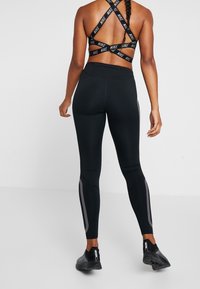 Sportovně založená žena stojí, má na sobě černou sportovní podprsenku Nike s kříženými ramínky a černé legíny s reflexními detaily, černé trenérky.