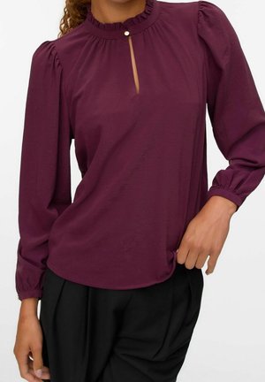 Frau trägt eine langärmelige, hochgeschlossene Pflaumenbluse mit kleinem Frontschlitz und Knopf, kombiniert mit schwarzen Hosen.