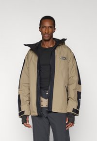 Μπεζ και μαύρο μπουφάν με κουκούλα, με λογότυπο στο στήθος, ελαστικές μανσέτες και άνετη γραμμή. Φορώνεται πάνω από μαύρο πουκάμισο.