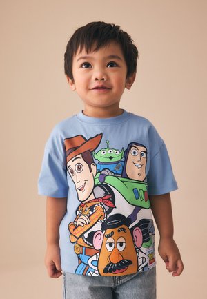 Ung dreng iført en blå Toy Story T-shirt med Woody, Buzz Lightyear, en alien og Hr. Kartoffelhovede mod en enkel beige baggrund.