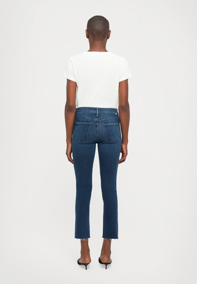 Persoon met kortgeknipt haar staat met de rug naar je toe, draagt een wit t-shirt, blauwe cropped jeans met ruwe zomen en zwarte haksandalen.