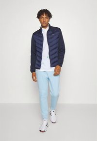 Casaco acolchoado azul-marinho com design matelassê, combinado com uma camisa polo branca e calças de treino azul claro. O modelo usa ténis brancos com detalhes escuros.