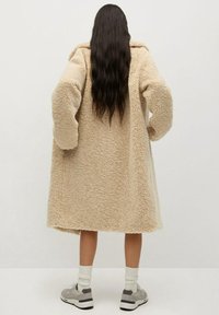 Vaaleanbeige, pörröinen, pitkä takki, jossa on ylikokoiset hihat ja teksturoitu kangas. Malli pukeutuu harmaisiin lenkkareihin ja valkoisiin sukkiin, kääntyneenä pois kamerasta.