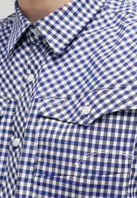 Blå och vit rutig skjorta med knappad krage, två bröstfickor och vita knappar; tillverkad av lätt bomullstyg.