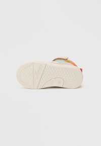 Värvikas laste spordijalats, millel on tekstureeritud beež tallad, velcro rihm ja mitmed sinise, oranži, punase ja rohelise suede aksessuaarid.