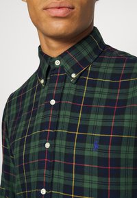 Green plaid shirt with blue, red, and yellow accents. Button-down collar and pearl buttons. Notable blue logo on the left chest.